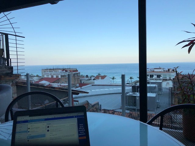 The top of my laptop in the bottom left corner, on a glass table, with the bay view in the background past red tile rooftops