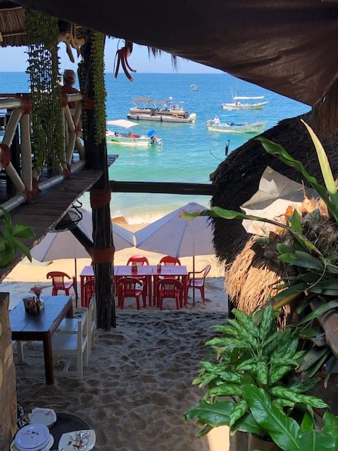 Looking down to the beach tables from inside the restaurant. Tables are nicely framed by the building on left and right, a sand floor, and small boats close to shore in the background.