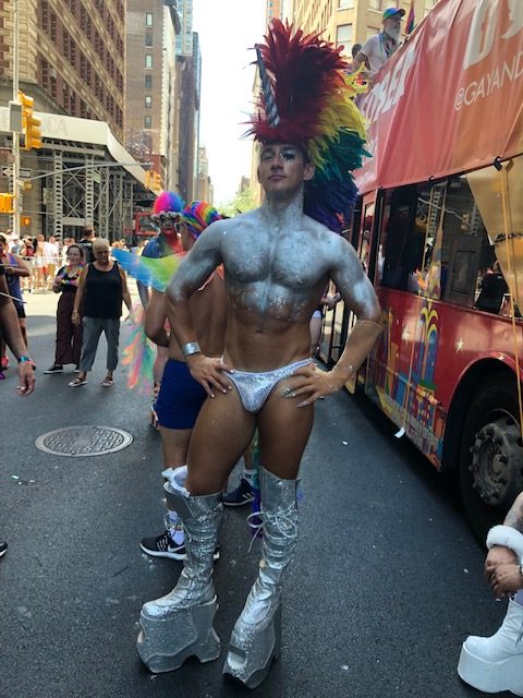 A gorgeous guy wearing only a shiny silver jockstrap, platform boots probably 10 inches high, silver glitter all over his torso and biceps, a rainbow feather mohawk headdress with a unicorn horn sticking out the front
