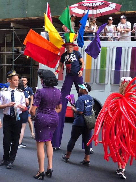 a guy on stilts with 6 flags sticking out of his bag in a peacock array, one for each of the rainbow colors