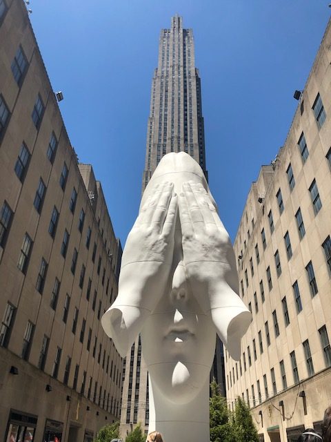 A 21 foot high sculpture of a girl's face with her hands - just hands, no arms - over her eyes, with Rockefeller Center behind it