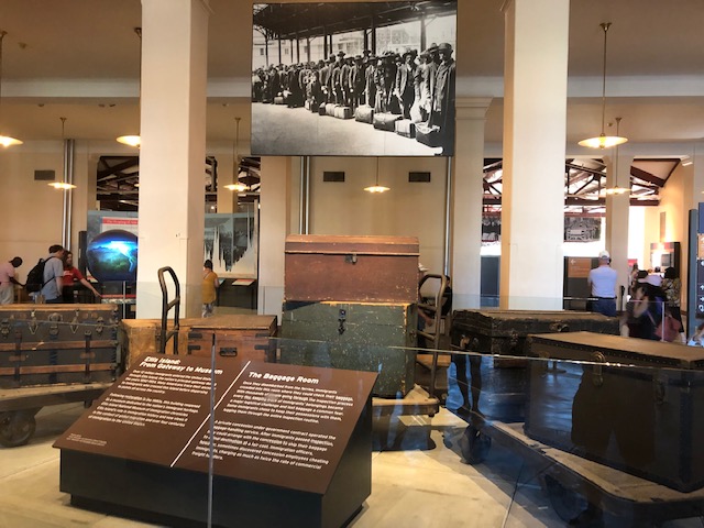 A display of trunks, probably 12 feet wide, with a black and white photo of immigrants lined up with their luggage above it