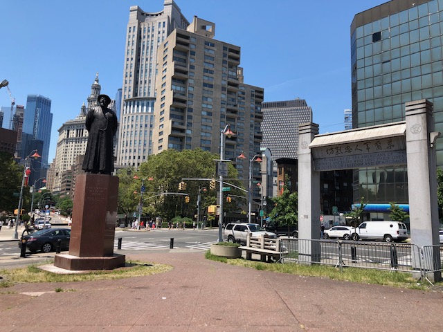 A statue of a man and an arch of sorts, at an intersection, tall buildings in the background