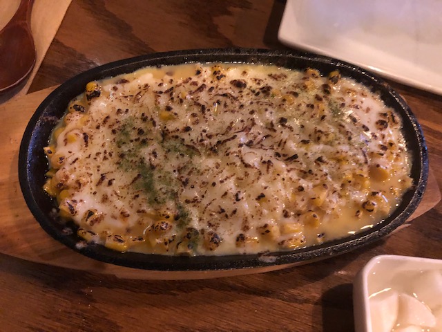 A small skillet with a dish called sizzling cheese corn; you can see yellow corn in sauce, white cheese, slightly burnt on top