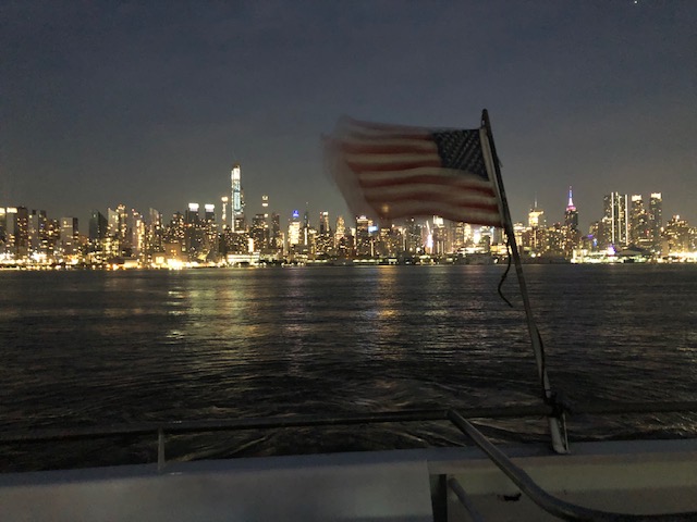 Midtown at night from the Hudson river, with a USA flag waving off the back of the boat