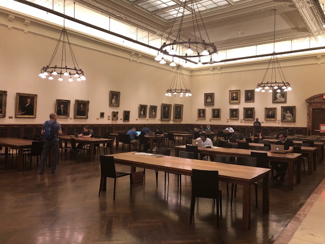 A huge room with long tables, people scattered throughout mostly working on laptops, walls covered in old painted portraits