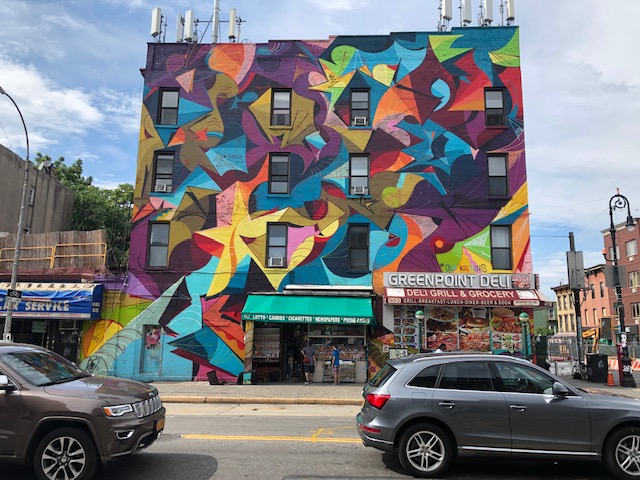A very modern mural covering a square building 4 stories high, no discernible design excetp for a yellos start bottom left. Other colors include lime green, purple, orange, sky blue. There's a deli at the right bottom corner.