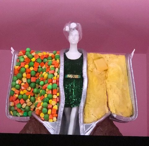 Mannequin in a simple sequined green short dress, with huge TV dinne trays hanging from the arms. One tray filled with traditional mixed vegetables: carrots, peas, corn; the other tray looks like cornbread with a slice of butter on top