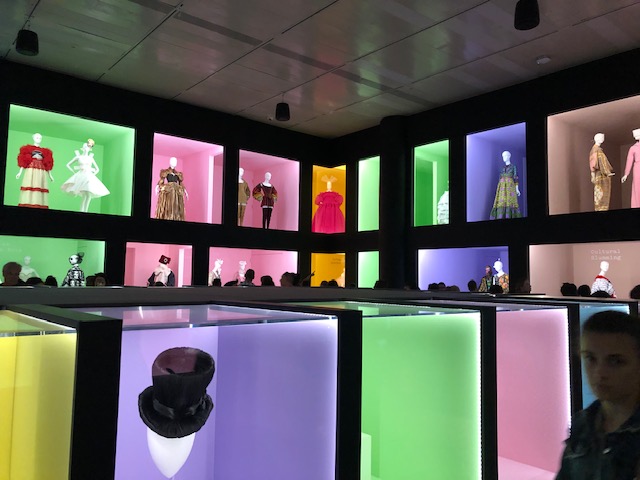 A large room with 2 levels of square windows, like department store sidewalk windows. Walls are black and room is unlit except for the light coming from the windows. 1-3 Mannequins in each window dressed in wide array of camp