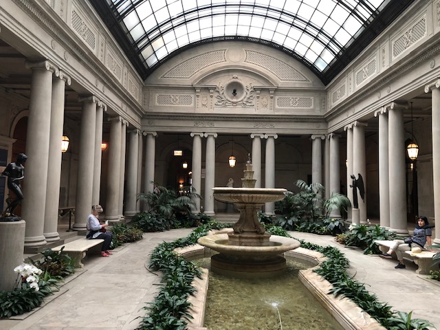 Atrium courtyard in the center. Columns along the edges, fountain in the middle, park benches, plants, statues