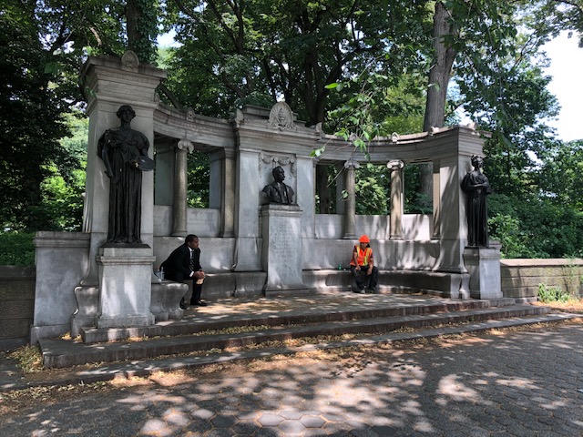 long curved bench along the sidewalk against columns, with a statue on either end and bust in the middle