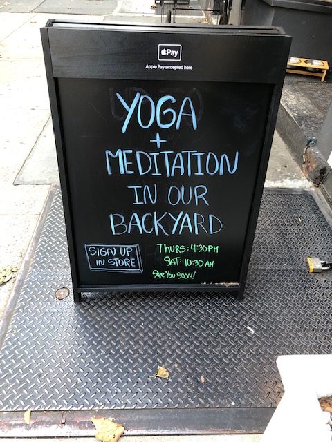 Sandwich board on the sidewalk that says "yoga and meditation in our backyard" with a schedule.