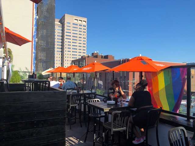 Small tables against glass wall at end of the rooftop with orange umbrellas, and rainbow flag draped over the outside of the glass