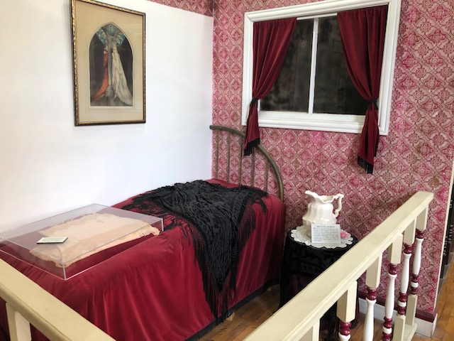 a small bed with a red satin spread, with a night table next to it, white urn on top