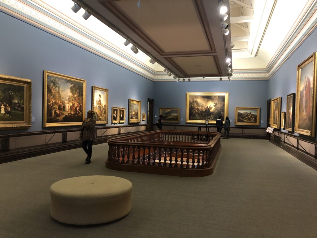 Large gallery of mostly very large paintings along the perimeter, with the middle open to looking down into the ballroom over a mahogany rail. 