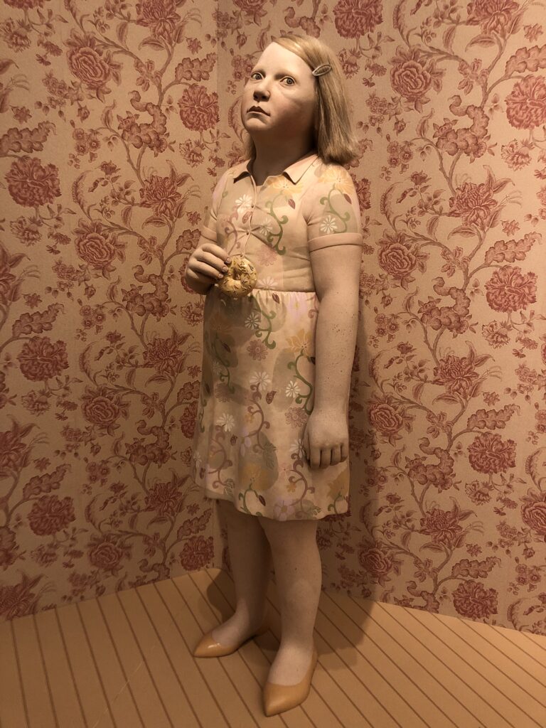 A stoneware  statue of a young girl, maybe 10, wearing a dress. She has hair, pulled back by a barrette to one side. She is standing in a corner holding a doughnut with a look of terror on her face. The walls behind her are papered in flowers. The entire thing is done essentially in varying shades of peach, muted reds and orange, with a tiny big of pale green in her dress connecting the flowers in the print. 