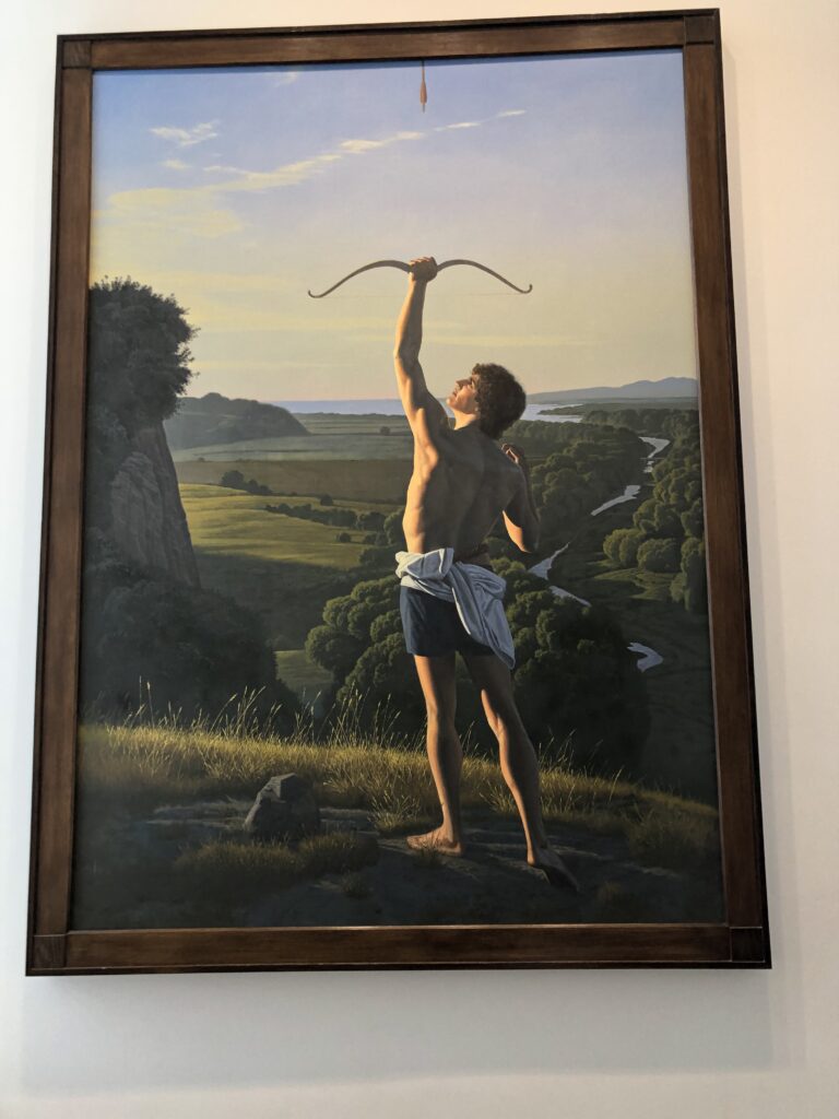 A shirtless man, mid-20s maybe, with his shirt tied around his shorts. His left arm is raised still holding the bow he just released an arrow from, his right arm pulled back in a crook from the release. The bottom tip of the arrow is seen at the top of the painting. He's standing on a hill, with a grassy valley and a behind him on the left and a river flowing through trees on the right.