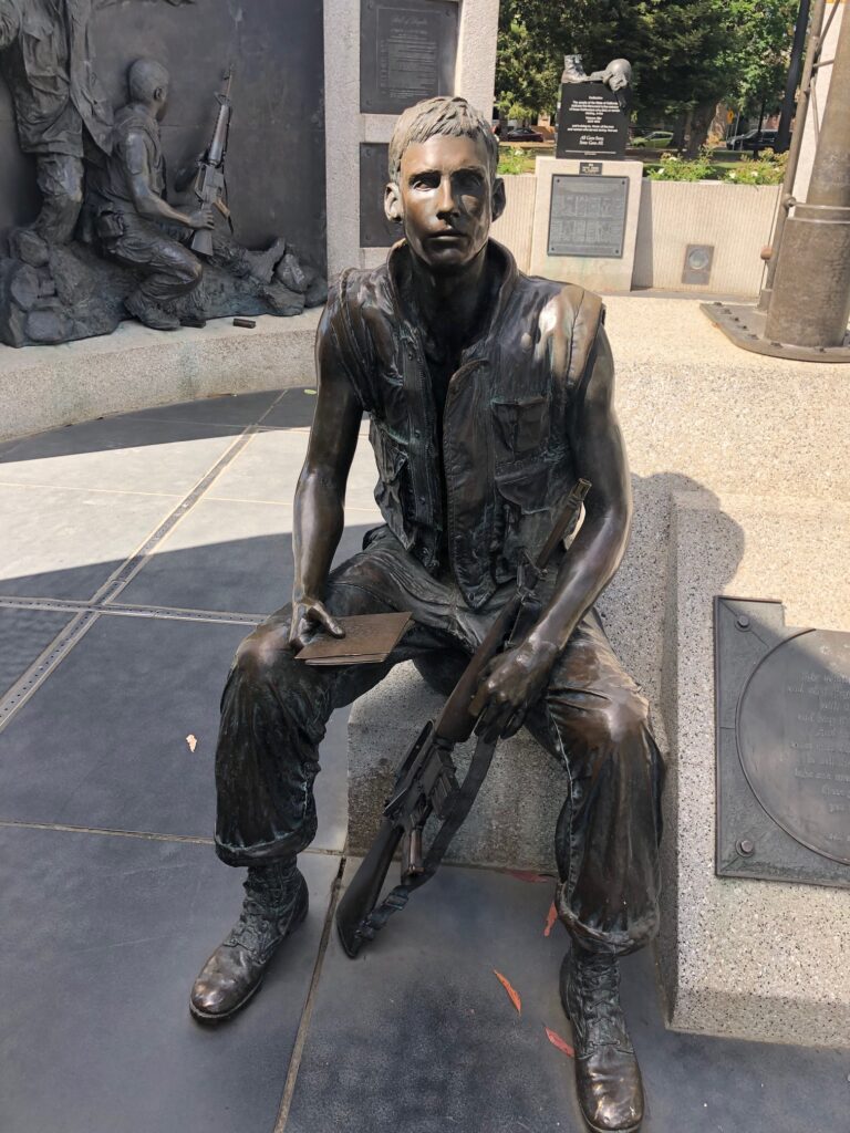 Sculpture of a young man sitting down holding a rifle. 