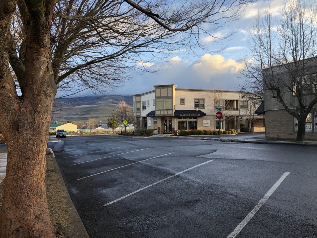 Empty parking spaces and street in foreground, buildings mid way, hills and sky in the background. 