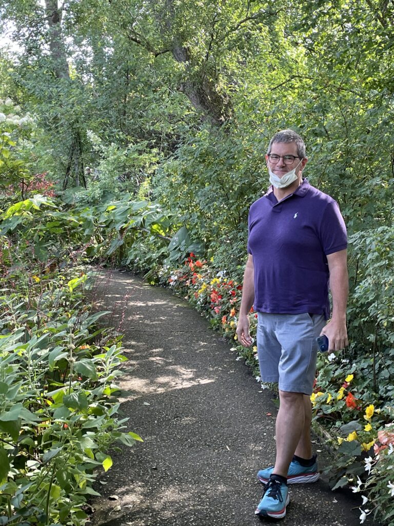 Me wearing a purple polo, gray shorts, and a big smile, walking along one of the paths with flowers on either side. 