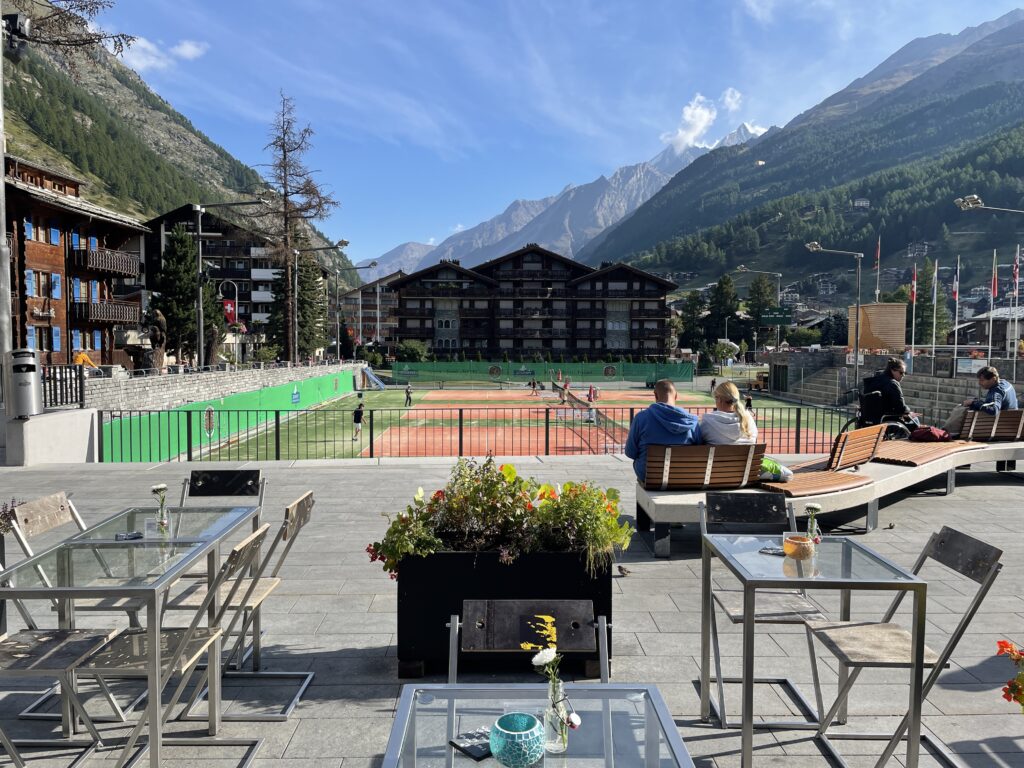 tables and chairs on a wide sidewalk, tennis courts behind that, hotels and mountains behind that 