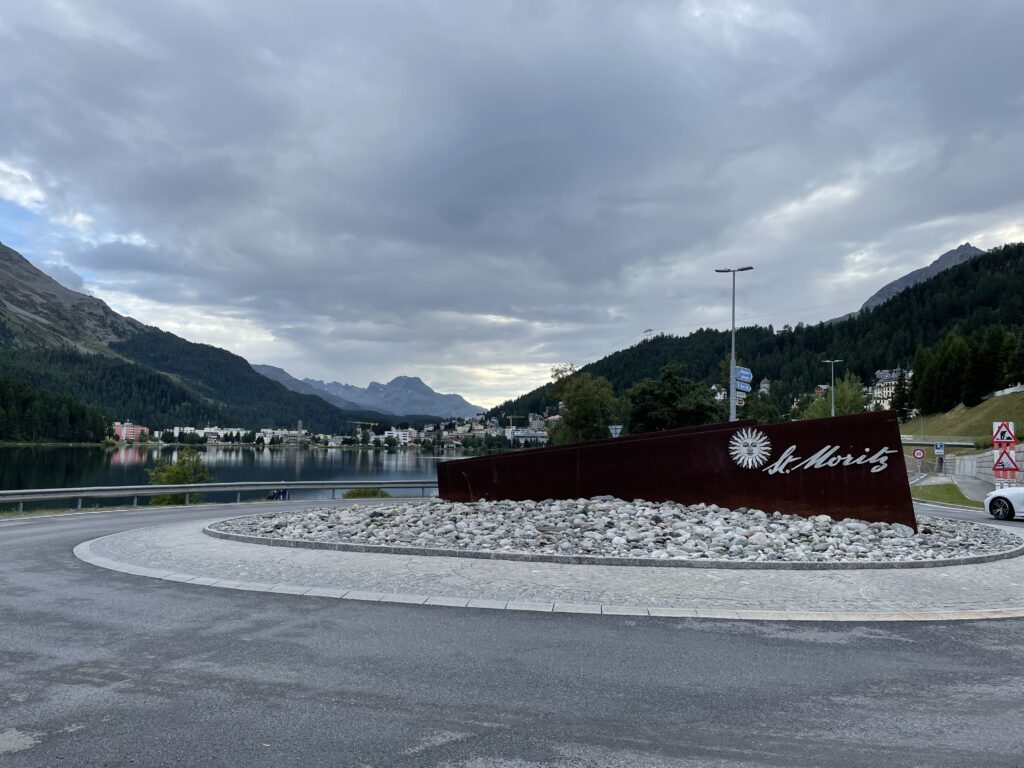 Decorative St Moritz sign in the middle of a roundabout next to the lake, with mountains and town in the background. 
